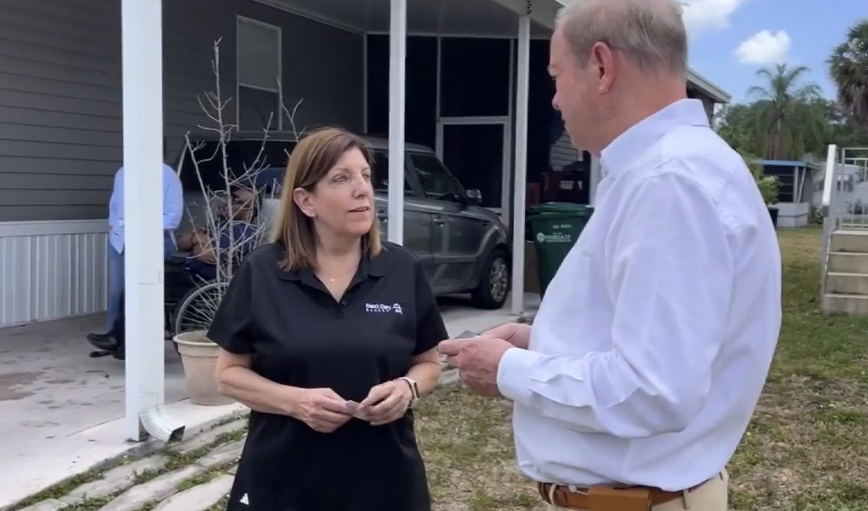A woman in a black polo shirt talking to a man in a white collared shirt outside a gray house.